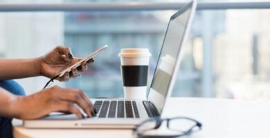 Close-up of hands using a laptop and phone with coffee on a modern office desk.